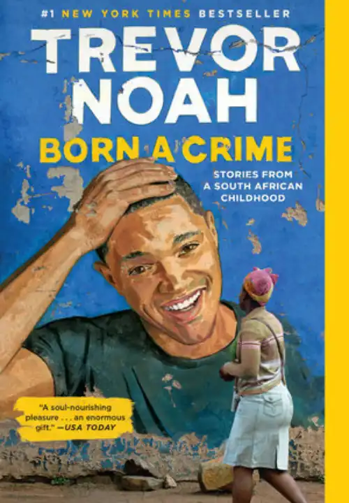 Book Cover of Born a Crime. A blue wall with paint peeling has a painted mural of Trevor Noah. Trevor is painted from the chest up. He is wearing a green t-shirt and has one hand placed on his head. Trevor looks straight ahead and is smiling, showing his teeth. A black woman faces the mural as she walks past it. The floor is dirt. The title of the book is painted above the mural.