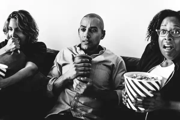 A black & white photo of a trio of friends watching a movie together. The first friend is biting their fist. The second friend is clutching a coffee cup and the third friend is holding a bucket of popcorn and grimacing.